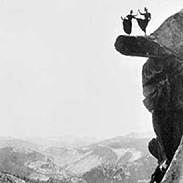 Two women in long skirts dance on a rock outcrop high above the ground